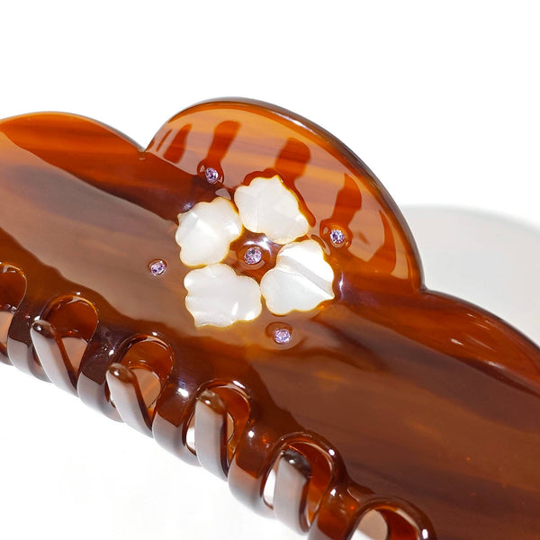 A detailed view of a glossy amber-brown hair claw clip with a scalloped edge, featuring a white floral embellishment and small purple rhinestone accents, shown on a white background.