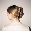 A woman shown from the back wearing a dark brown glossy tortoiseshell hair claw clip to style her blonde hair into a low bun, against a plain off-white wall.