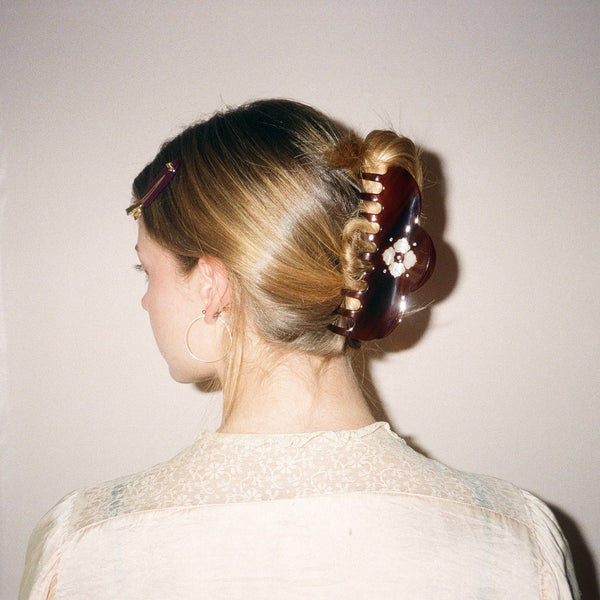 A woman shown from the back wearing a dark brown glossy tortoiseshell hair claw clip to style her blonde hair into a low bun, against a plain off-white wall.