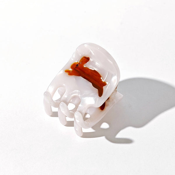 A small white iridescent hair claw with an amber brown rabbit accented with a tiny amethyst crystal eye, photographed on a white backdrop with a soft shadow.