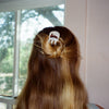 A person viewed from behind with long light brown hair, holding the hair half-up in place with a white hair clip decorated with a small amber rabbit. The background shows a window with a blurry outdoor view and a light-colored wall.