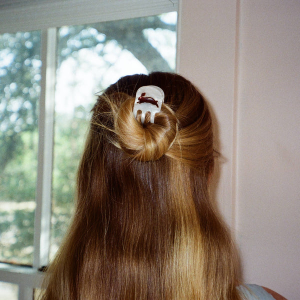 A person viewed from behind with long light brown hair, holding the hair half-up in place with a white hair clip decorated with a small amber rabbit. The background shows a window with a blurry outdoor view and a light-colored wall.