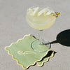 A coupe glass filled with a pale yellow cocktail sits on a light green scalloped coaster with gold trim and a gold monogram, placed on a neutral gray surface with a hard shadow.