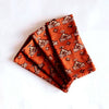 A stack of four fabric napkins in a rich orange shade with off-white and black patterns against a white backdrop.
