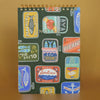 A top spiral-bound notebook with a dark green cover decorated with colorful vintage-style fish can labels with a gold embossed 'NOTES', photographed against a warm tan background.