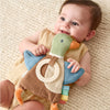 A baby lying on a textured cream blanket, smiling while holding a plush duck teether toy with a cream silicone teether ring and colorful rattle.