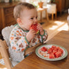 Baby wearing a woodland animals print bib, eating watermelon in a high chair with a plate of watermelon slices on a wooden table.