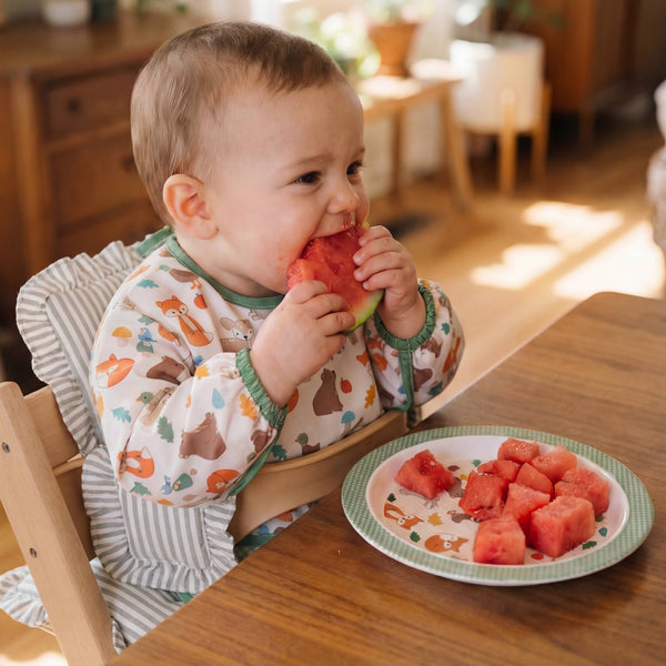 Baby wearing a woodland animals print bib, eating watermelon in a high chair with a plate of watermelon slices on a wooden table.