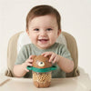 A baby sitting in a high chair holds a brown bear-shaped snack cup filled with cereal. The cup has a green rim and handle, and the baby wears a light green knit top against a plain light background.