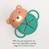 A children's snack cup with a bear-shaped lid in tan and green, shown on a light gray background with text below reading 'BPA-free and designed for the mess-free independent snacking stage from 6 months and up.'.
