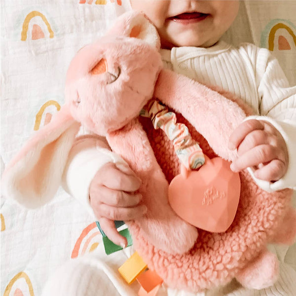 Baby holding a pink plush toy with a heart-shaped teether on a white blanket with rainbow patterns.