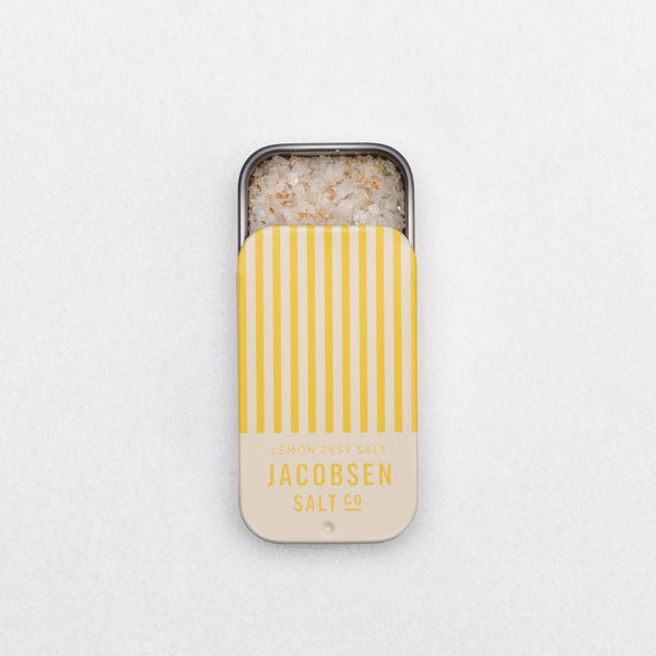 Small sliding tin container of lemon zest-infused sea salt with yellow striped lid on a white background