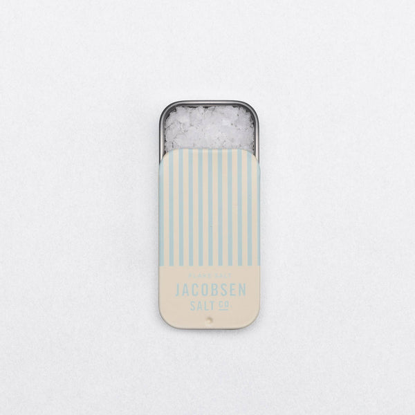 Metal slide tin of Jacobsen Salt Co pure flake sea salt with a blue and cream striped lid against a white background.