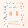 A pair of ballet shoes stud earrings displayed on a white JaxKelly brand card. The earrings are made of gold-toned metal with pink enamel detailing on the feet and bows.