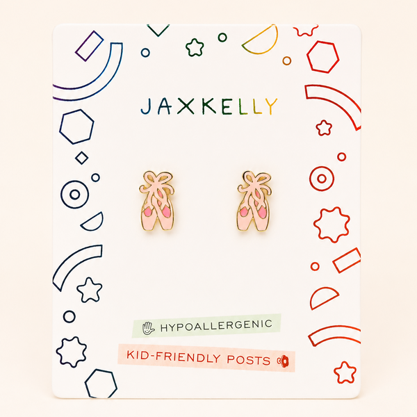A pair of ballet shoes stud earrings displayed on a white JaxKelly brand card. The earrings are made of gold-toned metal with pink enamel detailing on the feet and bows.