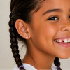 A close-up of a smiling young girl with dark braided hair wearing a small gold and pink ballet slippers stud earring against a light beige background.