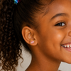 A close-up of a smiling young girl with curly brown hair in a ponytail, wearing a small football stud earring.