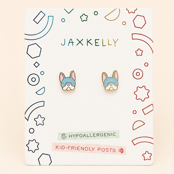 A pair of small French bulldog stud earrings displayed on a branded white backing card. The card shows the brand name JAXKELLY at the top, and the earrings feature pastel blue French bulldog faces with pink inner ears and cream/beige outlines.