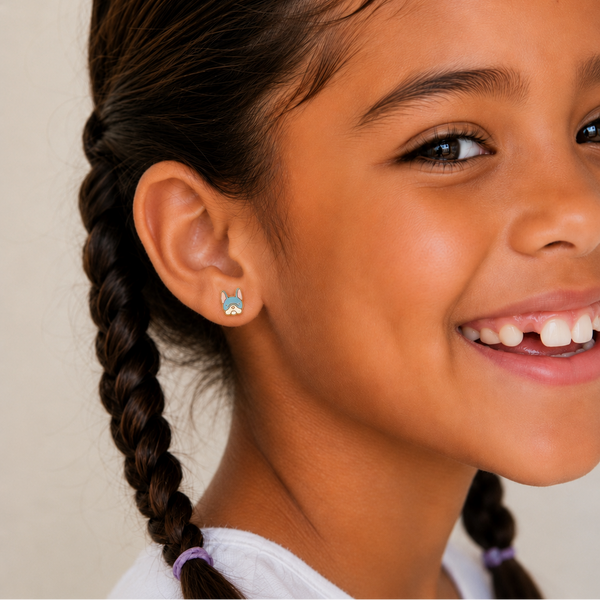 A close-up of a smiling young girl with dark braided hair wearing a small blue and white French bulldog-shaped stud earring.
