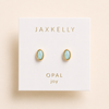 A pair of oval stud earrings with light blue fire opal centers and gold settings, displayed on a white Jaxkelly brand card labeled 'OPAL joy' against a warm beige background.