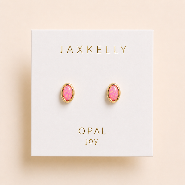 A pair of oval stud earrings with pink opal stones in gold bezel settings, mounted on a white JAXKELLY brand display card labeled 'OPAL joy' against a warm beige background.