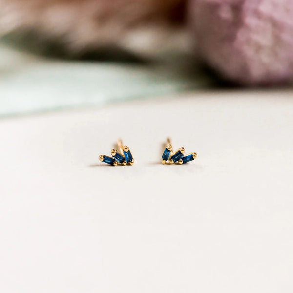 A pair of small gold stud earrings with an offset trio of tiny sapphire CZ stones, set against a blurred natural backdrop.