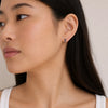 A person with long black hair wearing a white tank top looks to the side, showing a gold and sapphire trio stud earring in their ear. 