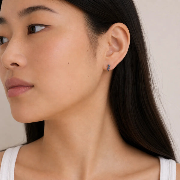 A person with long black hair wearing a white tank top looks to the side, showing a gold and sapphire trio stud earring in their ear. 