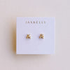 A white brand card with JAXKELLY' sits on a neutral backdrop holding a tiny pair of gold and white CZ stone double stack stud earrings.