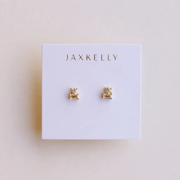 A white brand card with JAXKELLY' sits on a neutral backdrop holding a tiny pair of gold and white CZ stone double stack stud earrings.