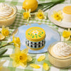 A tin of daffodil lip balm on a round mirror surrounded by yellow daffodils and sweet cream desserts on a green and white gingham picnic blanket. 