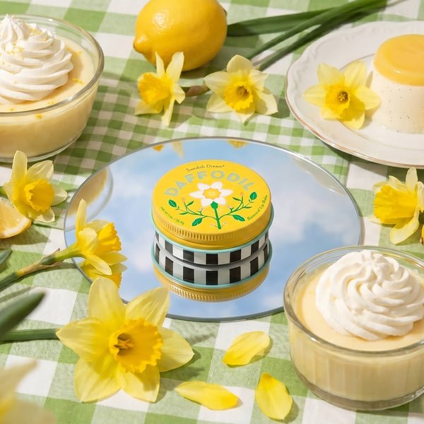 A tin of daffodil lip balm on a round mirror surrounded by yellow daffodils and sweet cream desserts on a green and white gingham picnic blanket. 