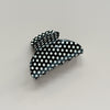 A glossy black hair claw clip with a white polka dot pattern, photographed against a neutral light gray background.