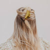 A blonde woman viewed from behind, wearing a scalloped yellow-green hair claw clip to secure a half-up style, with long wavy hair and a cream floral top dress against a plain light background.