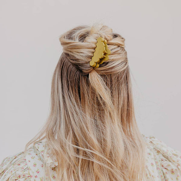 A blonde woman viewed from behind, wearing a scalloped yellow-green hair claw clip to secure a half-up style, with long wavy hair and a cream floral top dress against a plain light background.