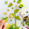Playful garden scene with a hand holding a magnifying glass, surrounded by small plants, flowers, and animals on a white background.