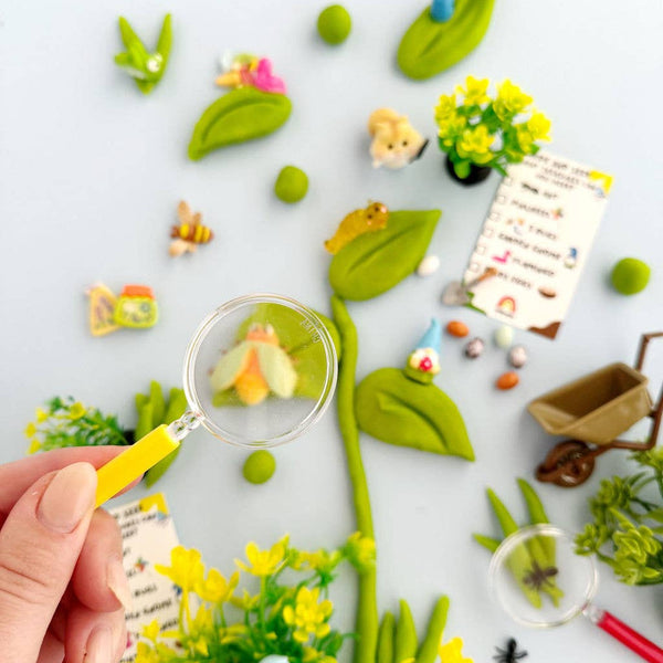 Playful garden scene with a hand holding a magnifying glass, surrounded by small plants, flowers, and animals on a white background.