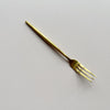 A single gold-colored metal appetizer fork with three tines, positioned diagonally on a textured off-white background.