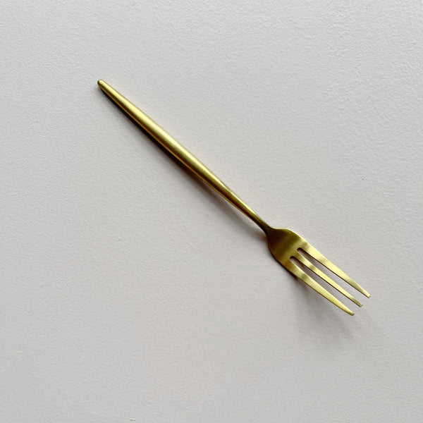 A single gold-colored metal appetizer fork with three tines, positioned diagonally on a textured off-white background.