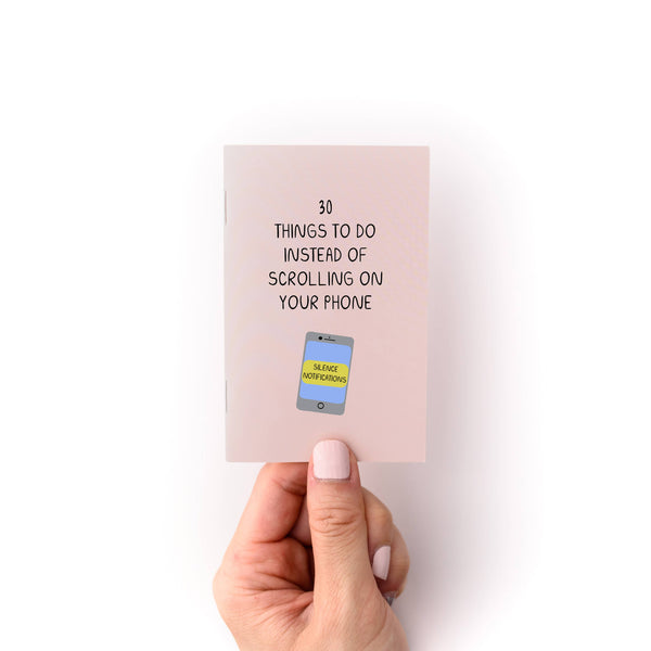 Hand holding a mini zine with text '30 Things to Do Instead of Scrolling on Your Phone' against a white background