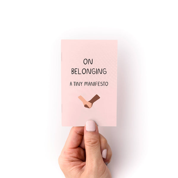 Hand holding a pink mini zine with text 'On Belonging: A Tiny Manifesto' on a white background