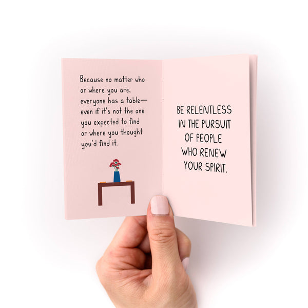 Hand holding a pink zine with text and a small illustration of a table and flowers on a white background