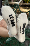 White socks with grippy black 'Le Bon Shoppe' branding worn by a person sitting on a green metal bench.