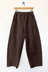 A pair of dark brown barrel-leg pants with an elastic waistband, hanging from a wooden hanger against a plain white wall.