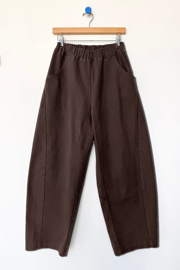A pair of dark brown barrel-leg pants with an elastic waistband, hanging from a wooden hanger against a plain white wall.