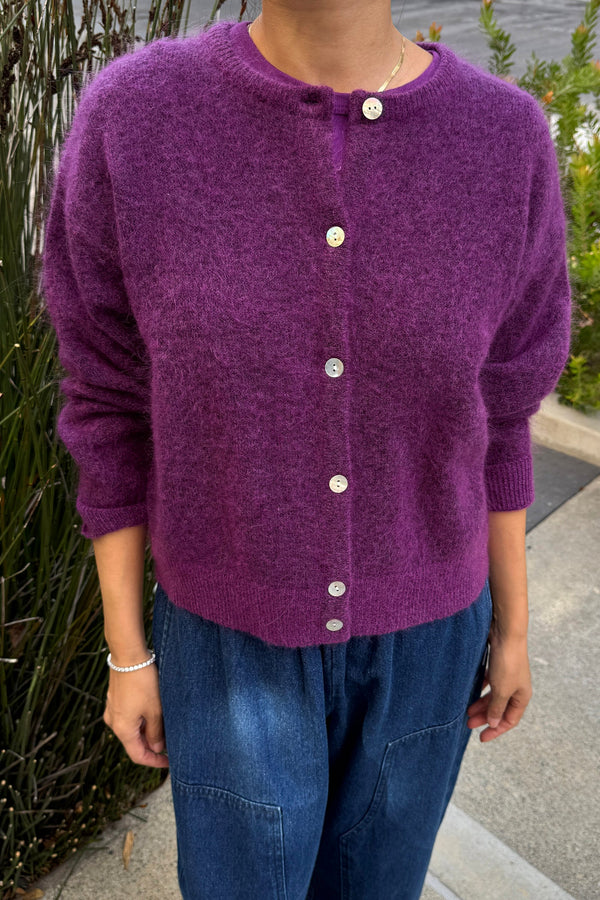A person wearing blue jeans and a rich purple fuzzy cardigan while standing on a sidewalk.