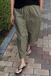 A person wearing army green elastic waist pants cuffed at the ankles with a black shirt and flip flops, walking along a cobblestone path.