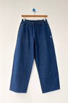 A pair of dark blue denim elastic waist pants on a wood hanger against a white wall.
