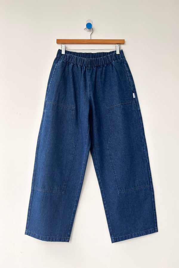 A pair of dark blue denim elastic waist pants on a wood hanger against a white wall.