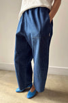A person wearing a pair of dark blue denim pants with a cream shirt and blue flats standing against a white wall.